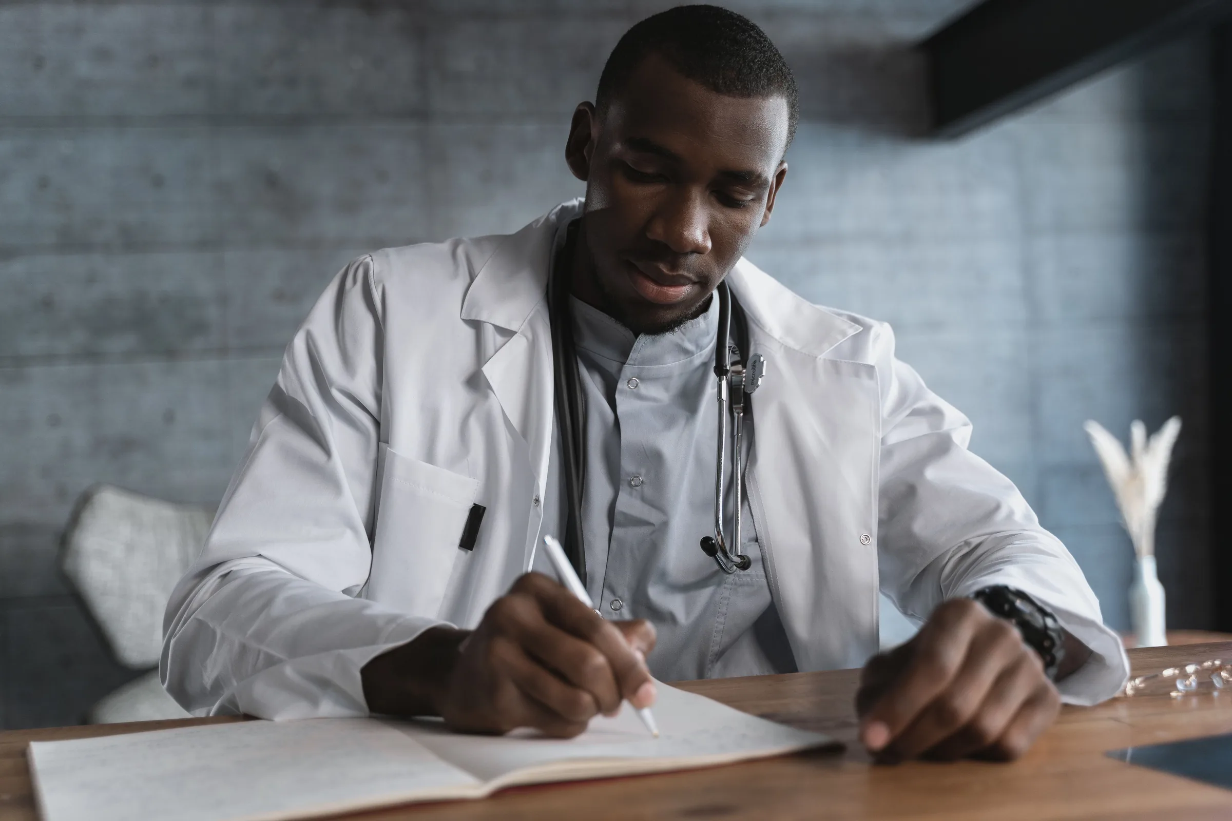 Doctor in white coat with stethoscope writing notes at desk in medical office.