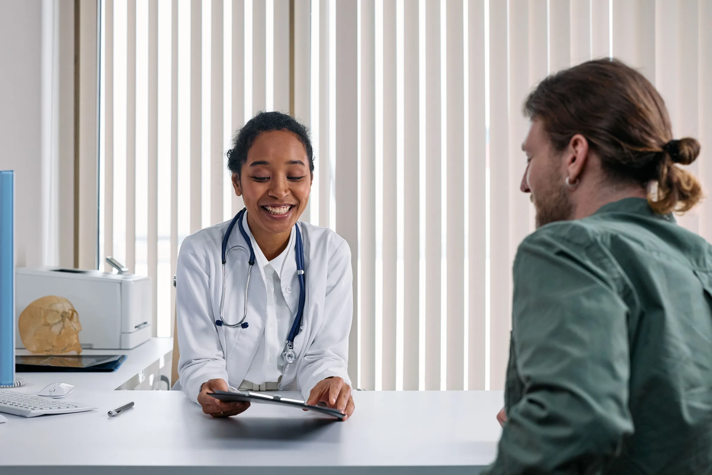 Smiling doctor in white coat with stethoscope consults with male patient at desk.